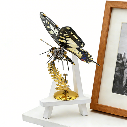 A stunning steampunk puzzle, a majestic mechanical swallowtail butterfly with intricate black, yellow, and white wings, beautifully displayed on a golden stand next to a small white easel and framed art. This impressive metal model showcases exceptional mechanical steampunk design, making it an ideal 3D puzzle Metal Kit for hobbyists or a captivating steampunk mech decorative piece that blends art and engineering.