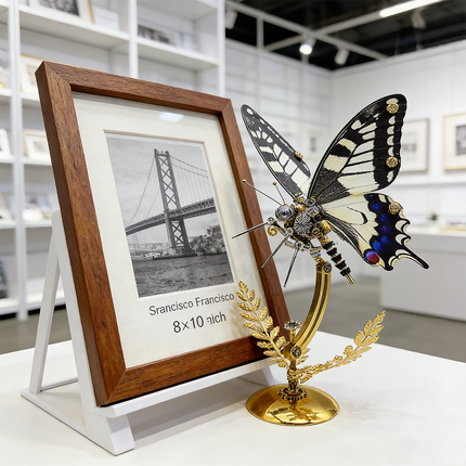 An artistic steampunk puzzle, a striking mechanical swallowtail butterfly with elegant black, white, and blue wings, elegantly displayed on a golden stand next to a framed picture of the Golden Gate Bridge in a modern gallery setting. This impressive metal model showcases sophisticated mechanical steampunk design, making it an ideal 3D puzzle Metal Kit or a captivating steampunk mech decorative piece that appeals to art lovers and travelers.
