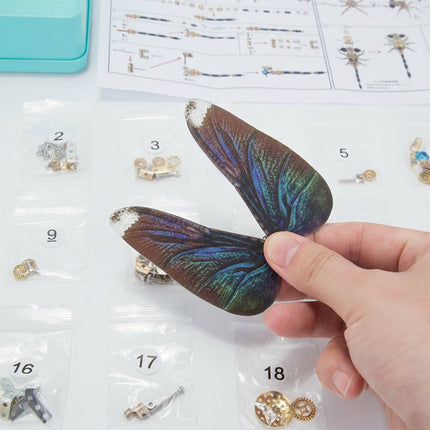 A hand holding a beautifully crafted, iridescent wing component for a mechanical dragonfly, amidst an array of numbered parts bags and assembly instructions for a steampunk puzzle. This close-up highlights the exquisite details and vibrant colors of the wing, a key feature of this metal model kit, demonstrating the meticulous design of this mechanical steampunk 3D puzzle Metal Kit or steampunk mech.