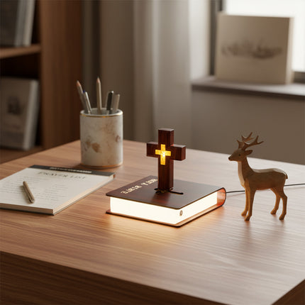 A floating cross lamp hovers above a luminous book-shaped base, placed on a home office desk adorned with a notebook, pen holder, and wooden deer figurine.