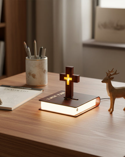 A floating cross lamp hovers above a luminous book-shaped base, placed on a home office desk adorned with a notebook, pen holder, and wooden deer figurine.
