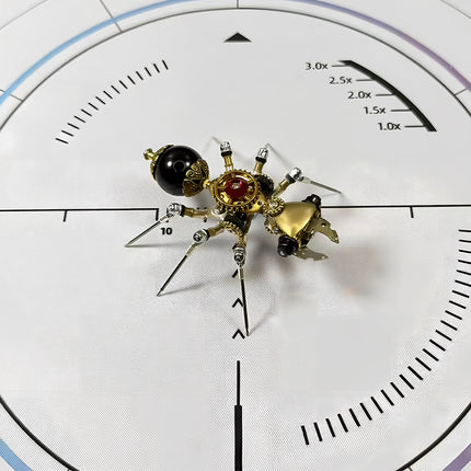  Overhead view of a mechanical steampunk ant 3D puzzle metal kit on a white measurement mat, showcasing its glossy black abdomen, golden head, and intricate mechanical details with a striking red accent. This steampunk puzzle is a remarkable piece.