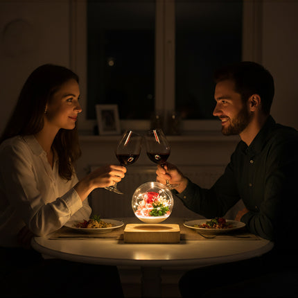 Floating anti-gravity glass orb containing preserved flowers, casting a warm glow from a wooden base between two people enjoying a romantic dinner at home