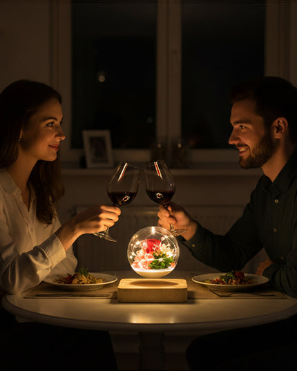 Floating anti-gravity glass orb containing preserved flowers, casting a warm glow from a wooden base between two people enjoying a romantic dinner at home
