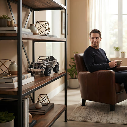 A man relaxing in a leather armchair in a cozy living room while looking at a finished 3D metal off-road vehicle model displayed on an industrial bookshelf.
