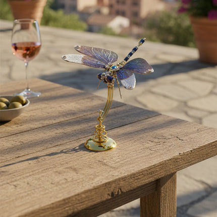 "Elegant assembled metal dragonfly model displayed on a golden stand, placed on a rustic wooden table outdoors with a glass of rosé and olives. A sophisticated maquette en metal, perfect as a decorative piece.