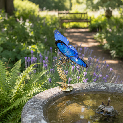 Vibrant blue butterfly metal model, a beautiful maquette en metal, perched on a stone bird bath in a serene garden with lavender and lush greenery. This exquisite metal model kit blends artistic craftsmanship with natural beauty.