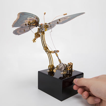 "Hand operating a steampunk butterfly metal model with teal and bronze wings, an interactive funny table sculpture. This DIY 3D puzzle metal kit provides engaging entertainment and a unique display.