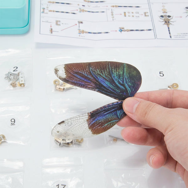 A close-up shot of a hand gently holding two beautifully crafted, iridescent wing components for a mechanical dragonfly, with various numbered parts bags and assembly instructions visible in the background. This image emphasizes the delicate and colorful nature of the wings, which are central to this steampunk puzzle metal model, making it an engaging 3D puzzle Metal Kit for constructing a unique mechanical steampunk creature or steampunk mech.