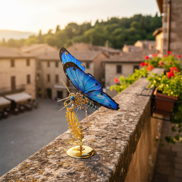 Stunning blue butterfly metal model, a beautiful maquette en metal, perched on a stone wall overlooking a picturesque European town at sunset. This intricate metal 3D puzzle makes a unique decorative gift.