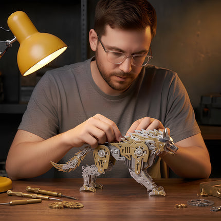 A man with glasses carefully assembling a gold and silver 3D metal mechanical wolf model at a workbench under a desk lamp.